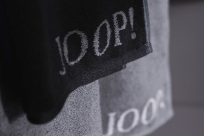 Produktbild: Seiflappen und Handtücher der Marke JOOP! in einem grauen Badezimmer.