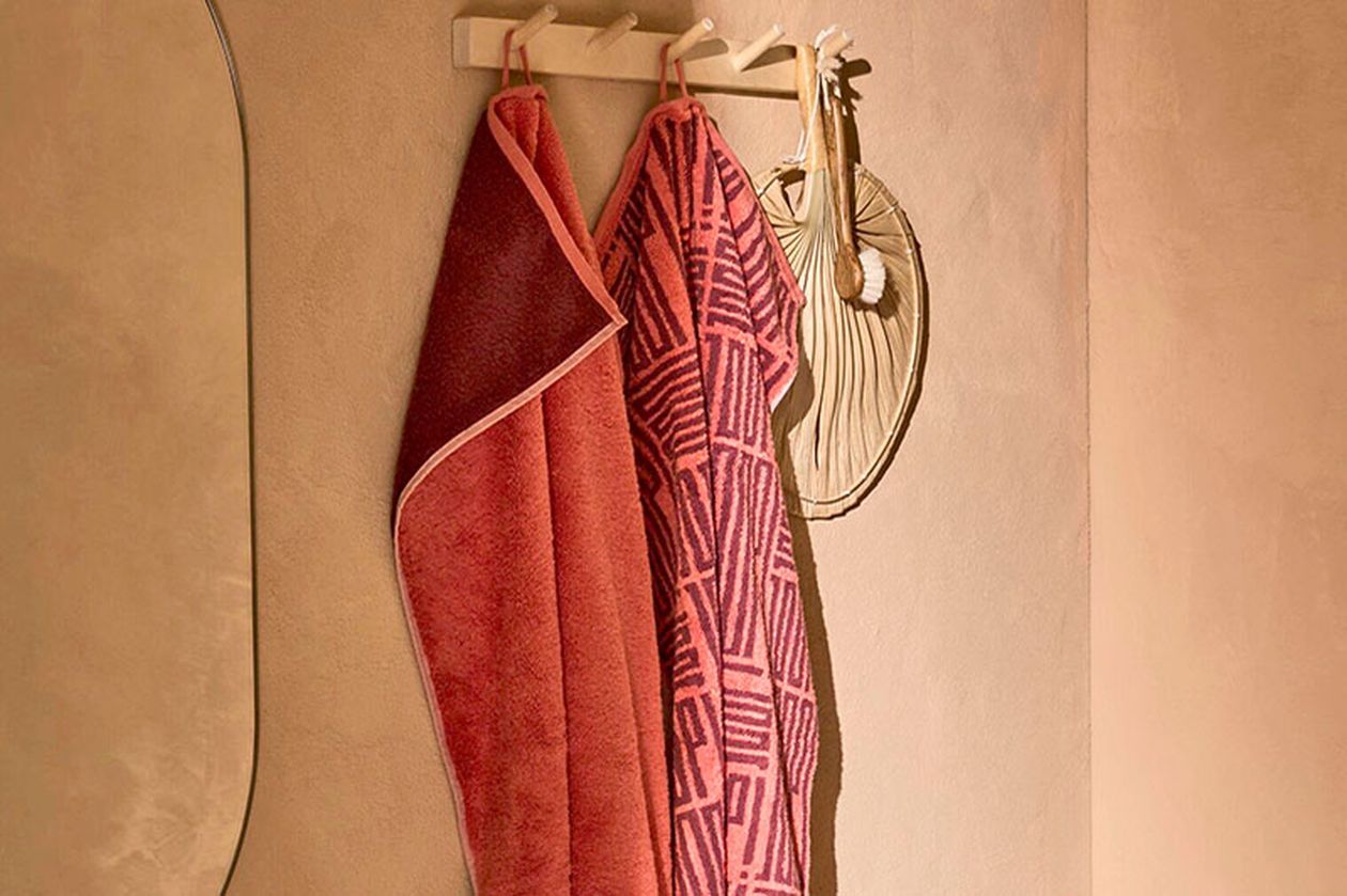 Zwei gemusterte rosa Handtücher hängen an Holzhaken an einer beigen Wand, daneben ein dekorativer Spiegel.