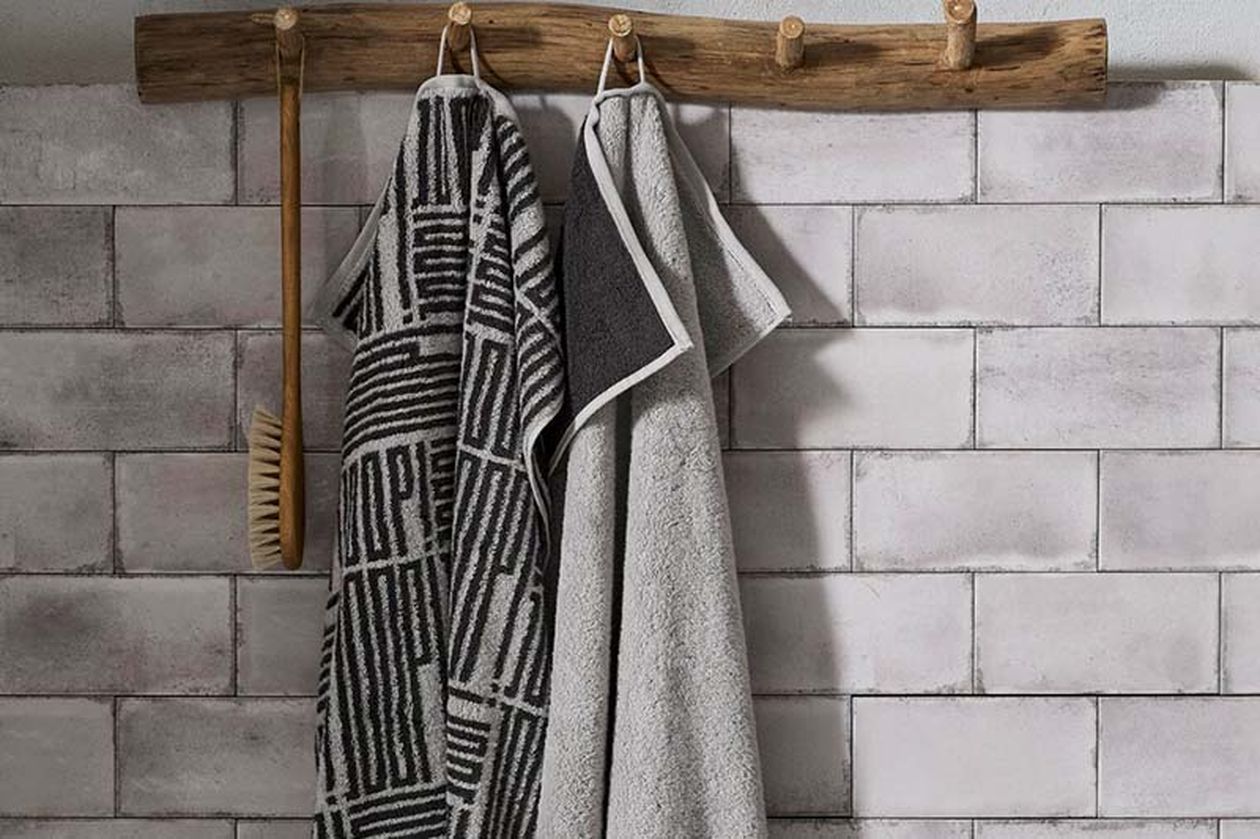 Zwei Handtücher, eines gemustert und eines einfarbig, hängen an einem rustikalen Holzgestell neben einer Bürste; im Hintergrund ist eine geflieste Wand zu sehen.
