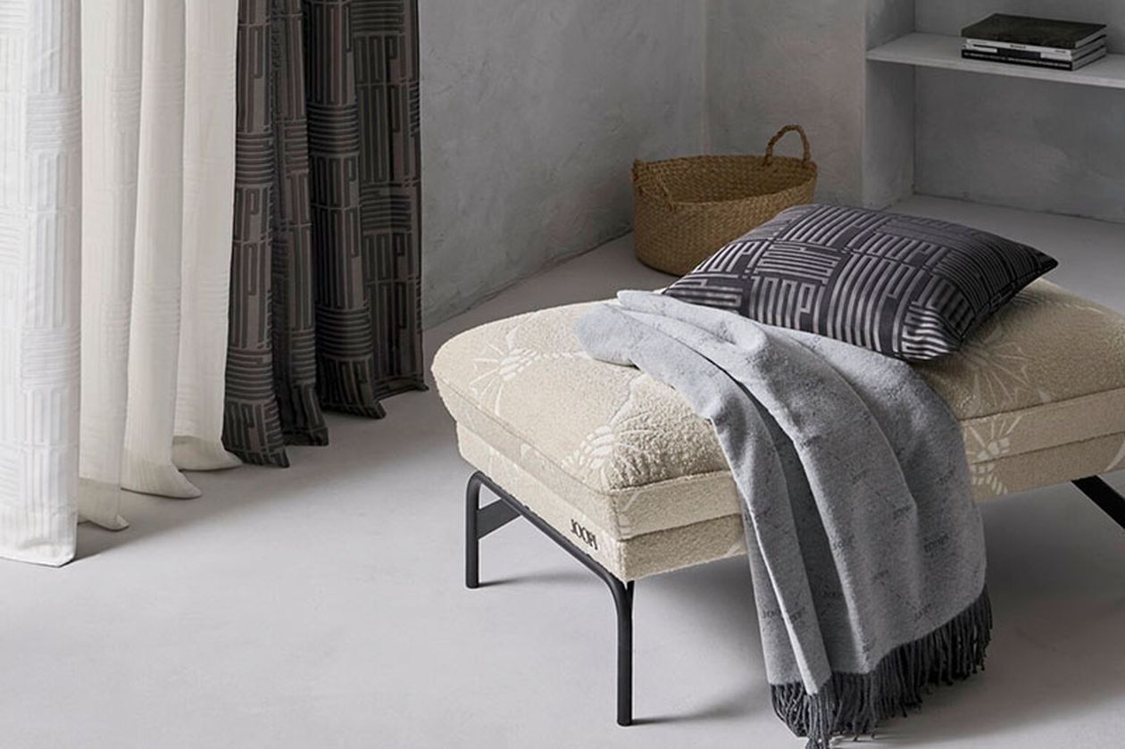 Modernes Zimmer mit beigefarbenem Hocker, grauer Decke, gemustertem Kissen und Korb. Vorhänge im Hintergrund; neutrale, minimalistische Einrichtung.