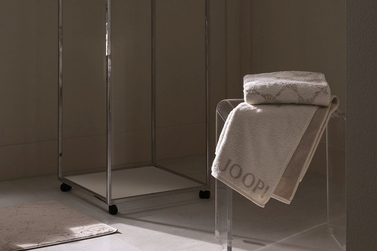 Produktbild: Seiflappen und Handtücher der Marke JOOP! auf einem Hocker in einem sandfarbenen Badezimmer.