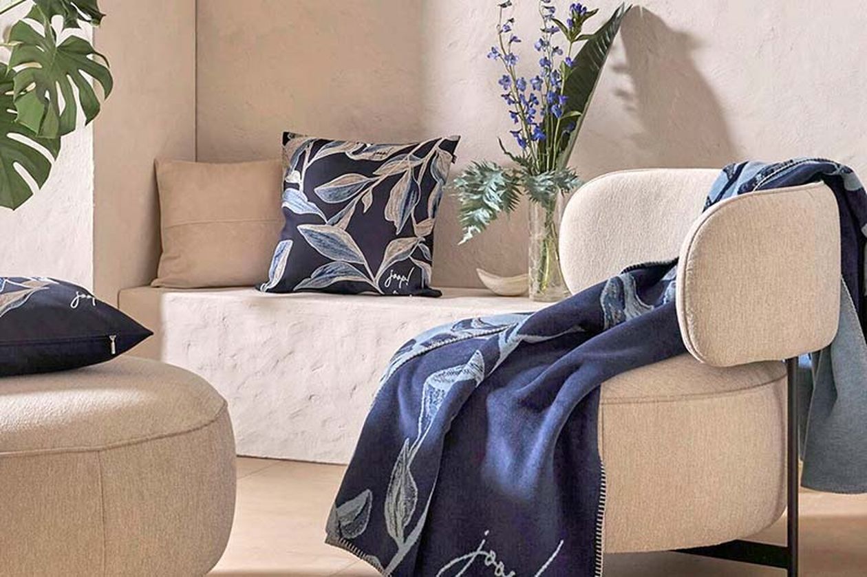 Modernes und ruhiges Zimmer mit beigefarbenem Sessel, blau gemusterten Kissen und passender Tagesdecke. Eine Vase mit Blumen schmückt die Steinbank.