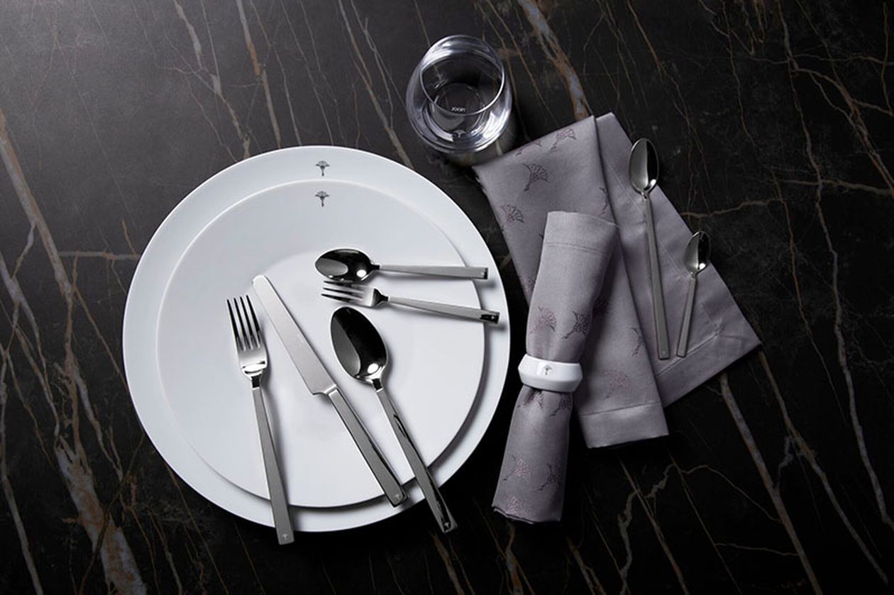 Gedeckter Tisch mit einem weißen Teller, silbernem Besteck, einem Glas und einer gefalteten grauen Serviette auf einer dunklen Marmoroberfläche.