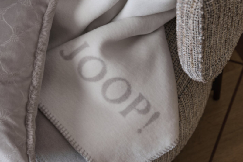 Produktbild: Kuscheldecke und Samtkissen der Marke JOOP! auf einem Sessel in einem hell gehaltenen Wohnzimmer.