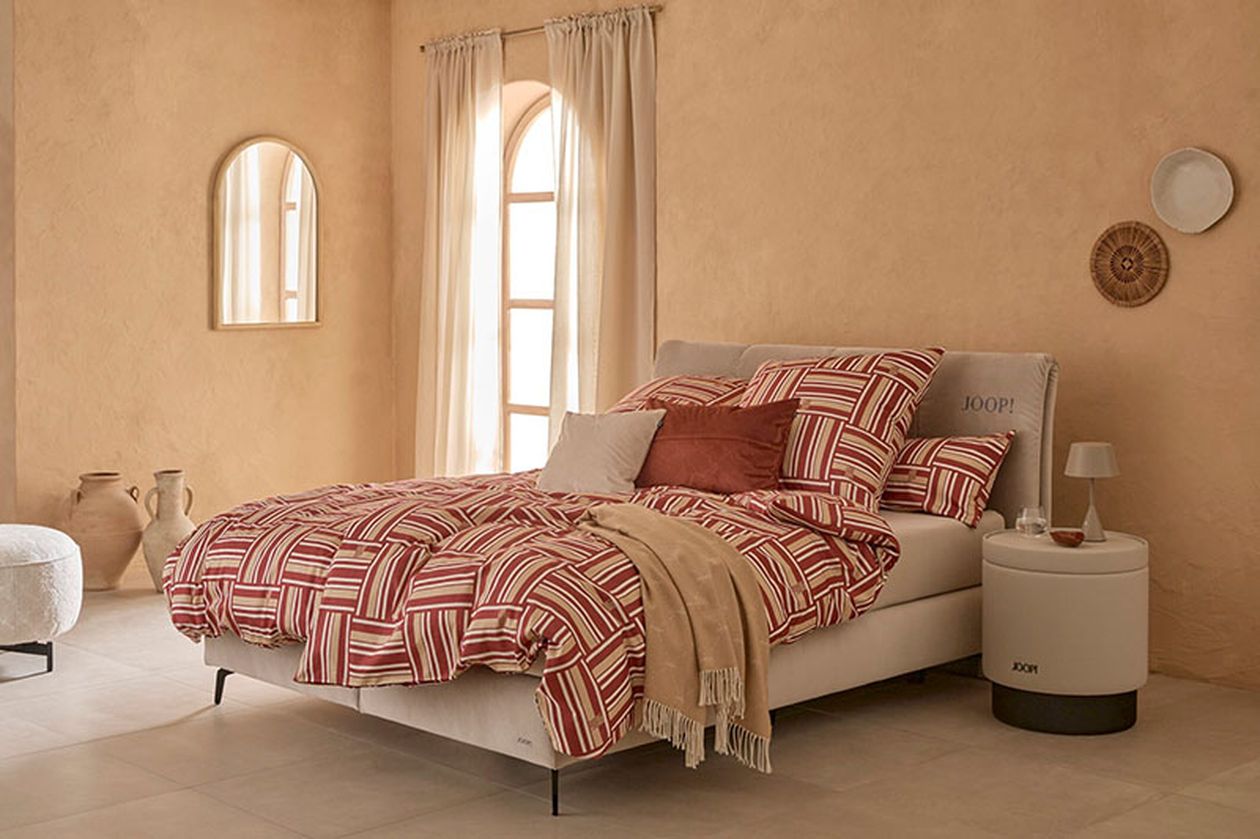 Schlafzimmer mit einem Bett, dessen Bettwäsche rot-weiß gestreift ist, passenden Kissen und einer beigefarbenen Decke. Neutrale Wände mit Spiegeln und Dekorationselementen.