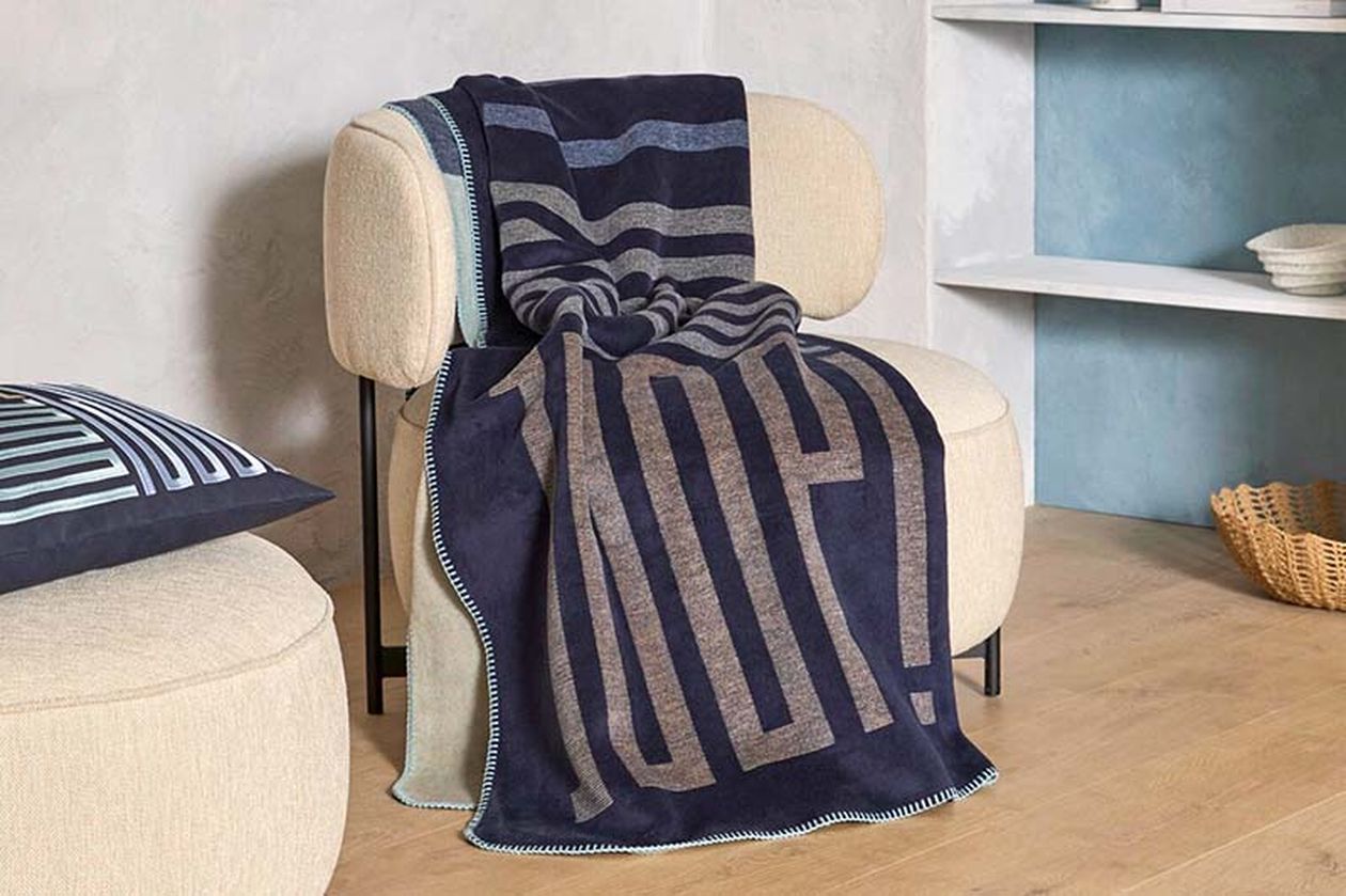 Ein beigefarbener Sessel, über den eine marineblau-grau gemusterte Decke drapiert ist, daneben ein passendes Kissen auf einem hellen Holzboden.