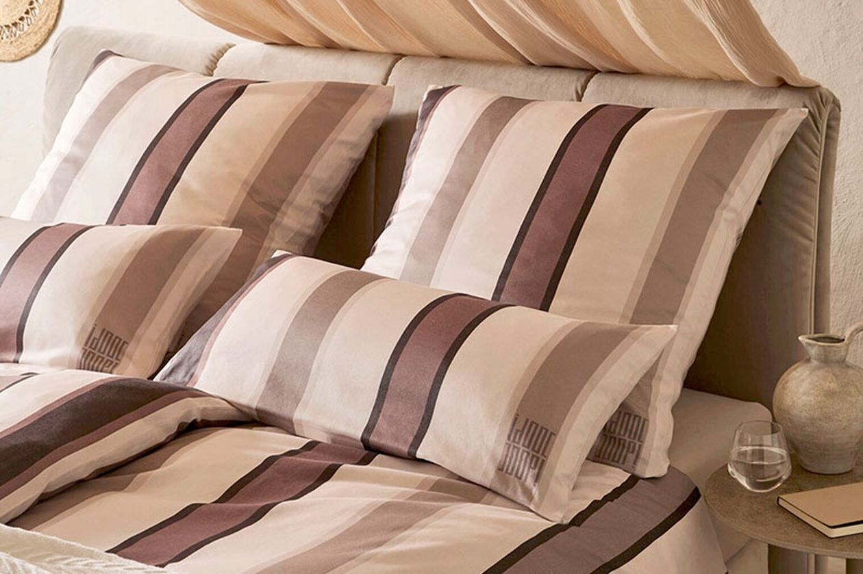 Ein ordentlich gemachtes Bett mit gestreiften beigen und braunen Kissen und Bettbezug, daneben ein kleiner Nachttisch mit einer Vase und einem Glas Wasser.