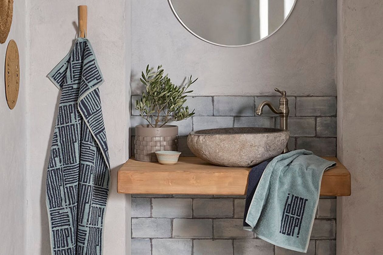 Minimalistisches Badezimmer mit rundem Spiegel, Steinwaschbecken, Topfpflanze und gemusterten Handtüchern auf einer Holzablage vor einer grau gefliesten Wand.