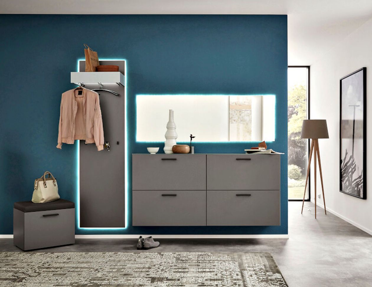 Moderner Flur mit blauer Akzentwand, beleuchtetem Spiegel, grauen Schränken, Garderobe mit einer rosa Jacke und einer Stehlampe am Fenster.