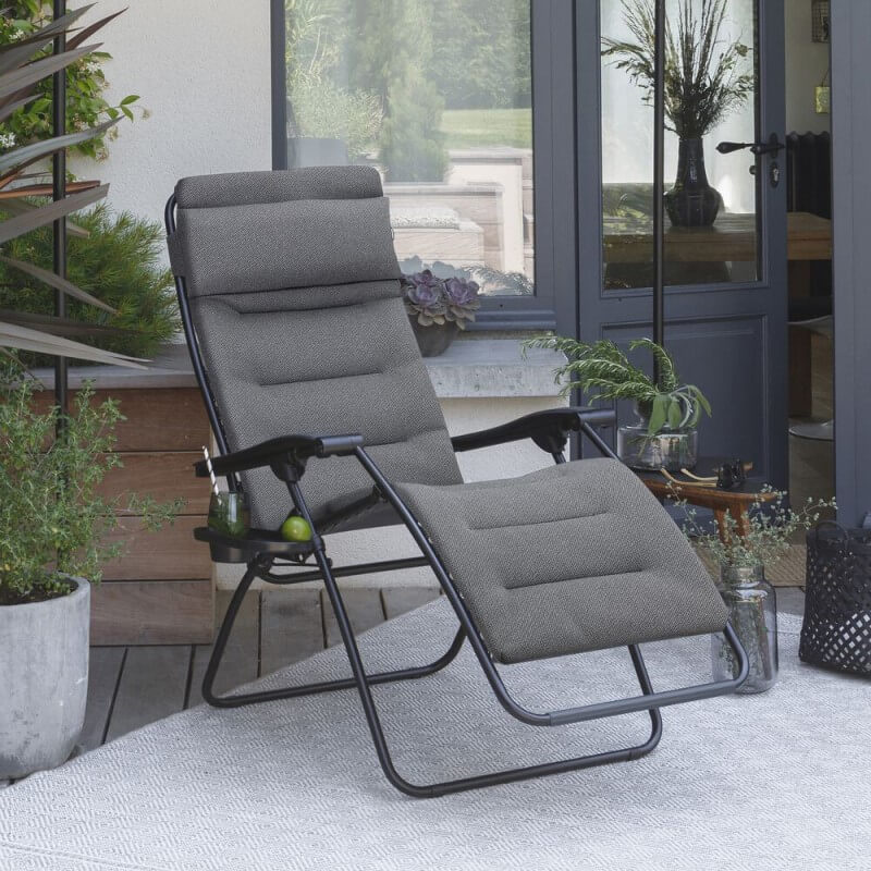 Ein grauer Liegesessel auf einer Terrasse, umgeben von Topfpflanzen und einer Glastür.