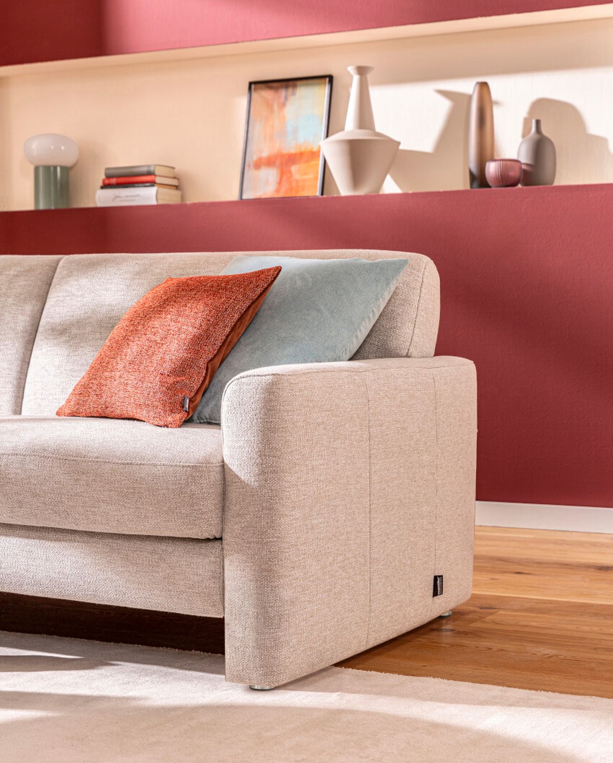 Beiges Sofa mit orangefarbenen und blauen Kissen in einem modernen Wohnzimmer. Rote Akzentwand mit Regalen, auf denen Dekoartikel ausgestellt sind, im Hintergrund.