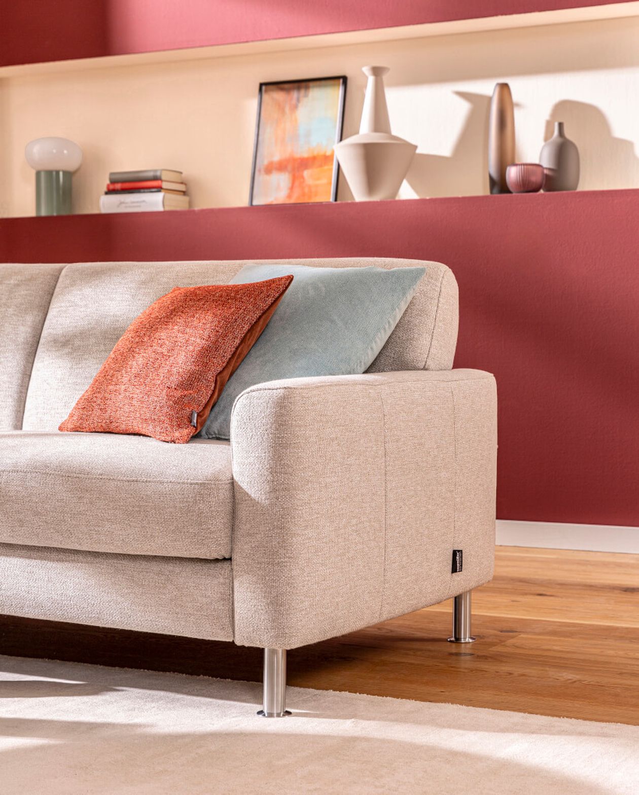 Beiges Sofa mit orangefarbenen und blauen Kissen in einem modernen Wohnzimmer. Rote Akzentwand mit Regalen, auf denen Dekoartikel ausgestellt sind, im Hintergrund.