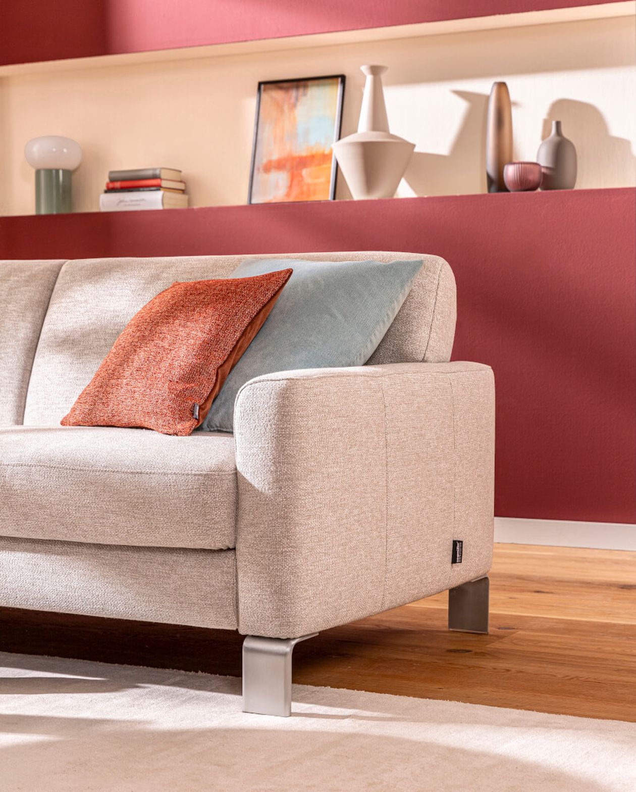 Beiges Sofa mit orangefarbenen und blauen Kissen in einem modernen Wohnzimmer. Rote Akzentwand mit Regalen, auf denen Dekoartikel ausgestellt sind, im Hintergrund.