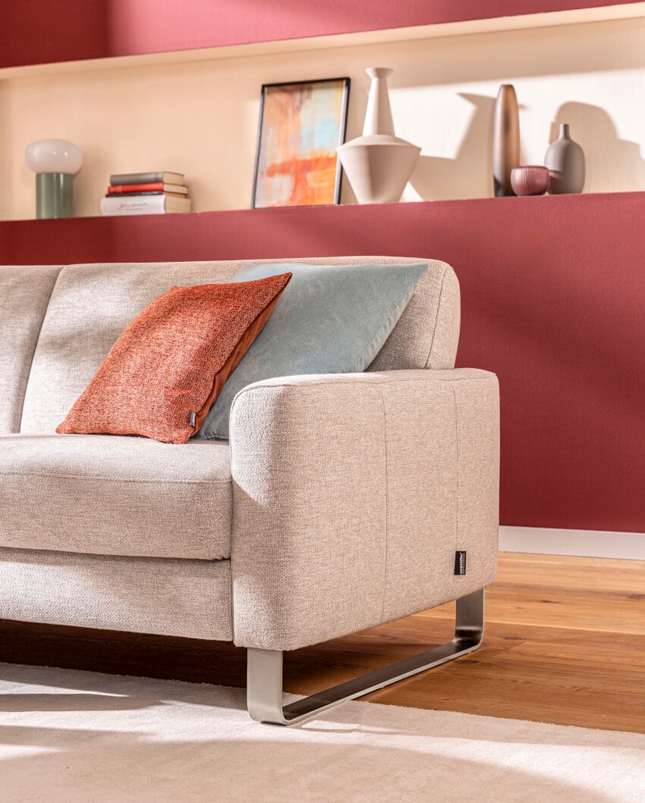 Beiges Sofa mit orangefarbenen und blauen Kissen in einem modernen Wohnzimmer. Rote Akzentwand mit Regalen, auf denen Dekoartikel ausgestellt sind, im Hintergrund.