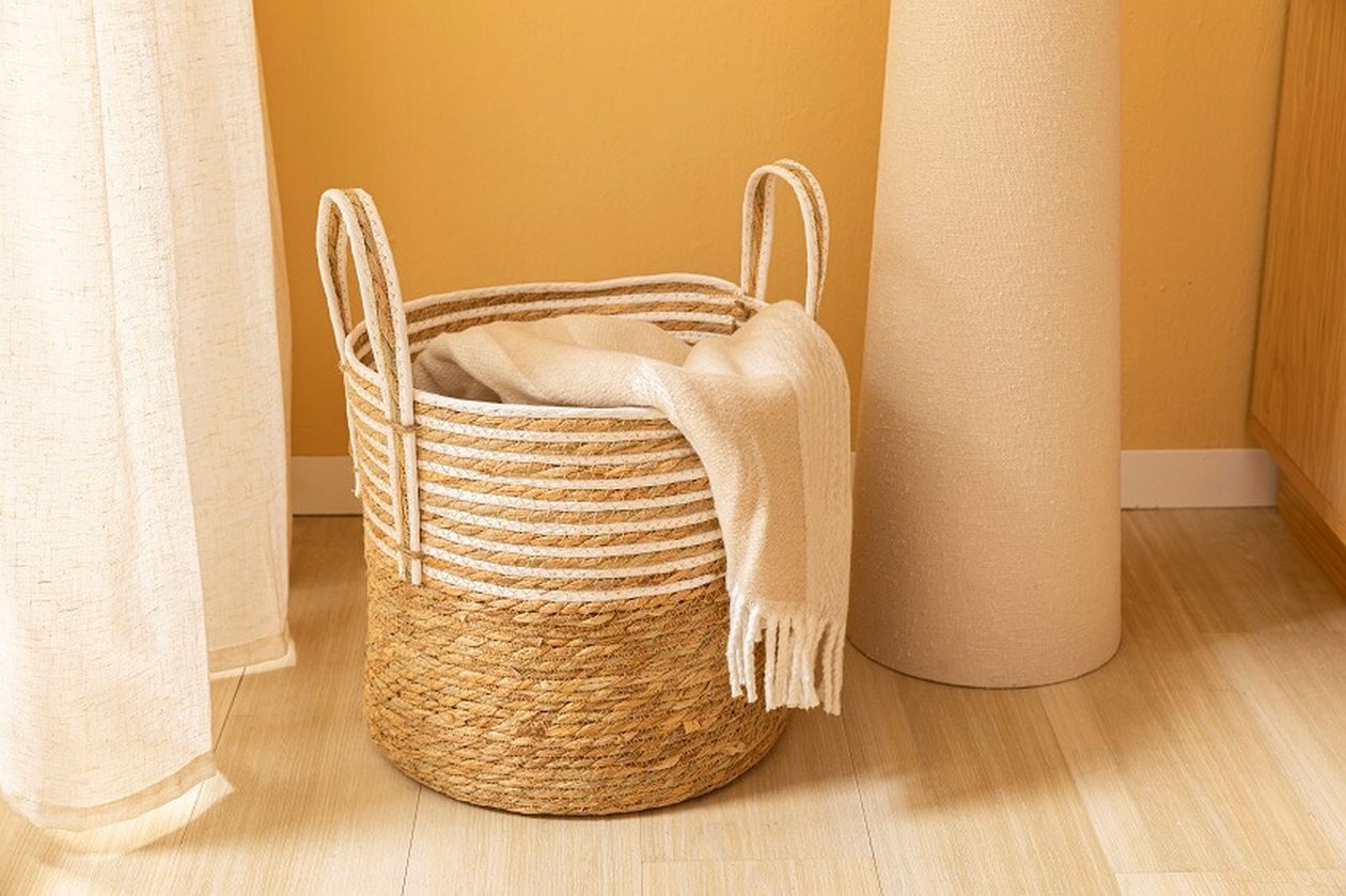 Eine beige Decke liegt über einem geflochtenen Korb mit Henkeln auf einem Holzboden vor einer warmgelben Wand in einem gemütlichen Interieur.