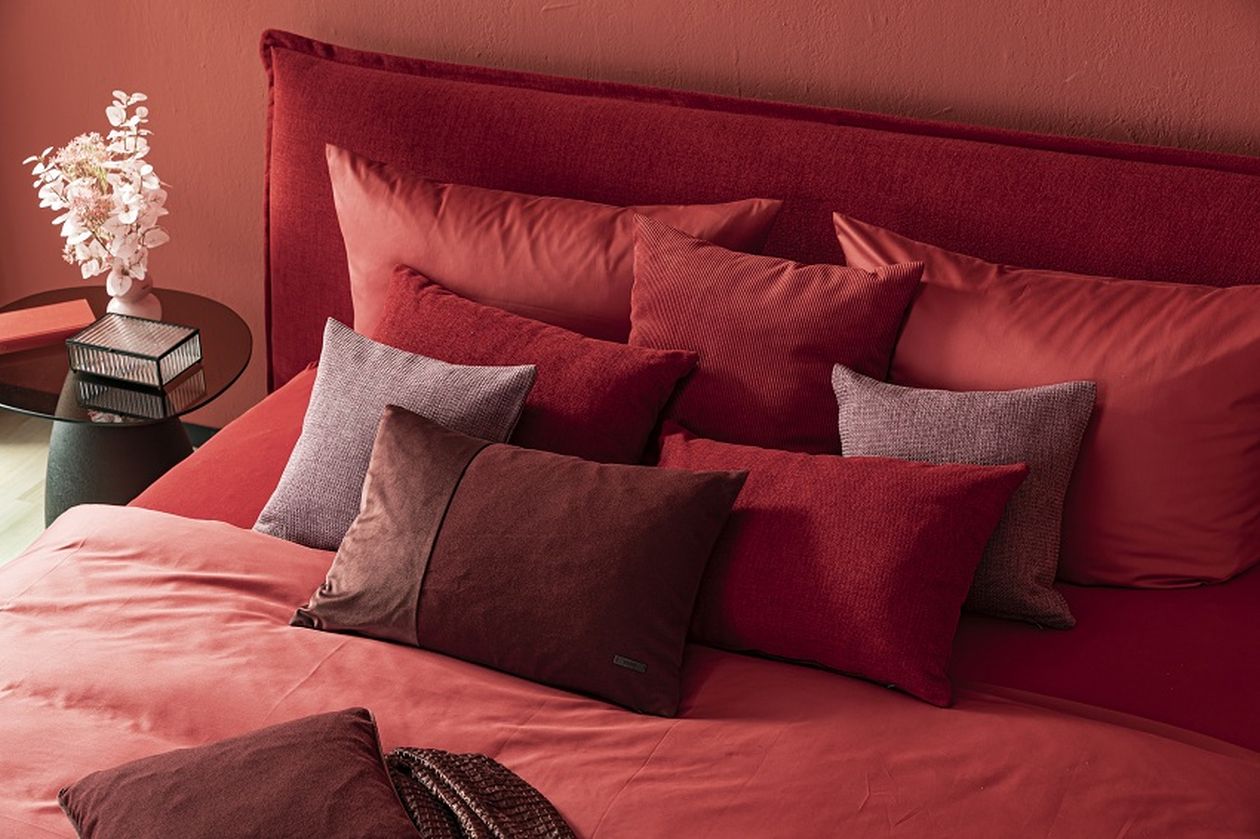 Rotes Bett mit verschiedenen Kissen in Rottönen, daneben ein runder Tisch mit Vase und Blumen, moderne und gemütliche Schlafzimmer-Dekoration.