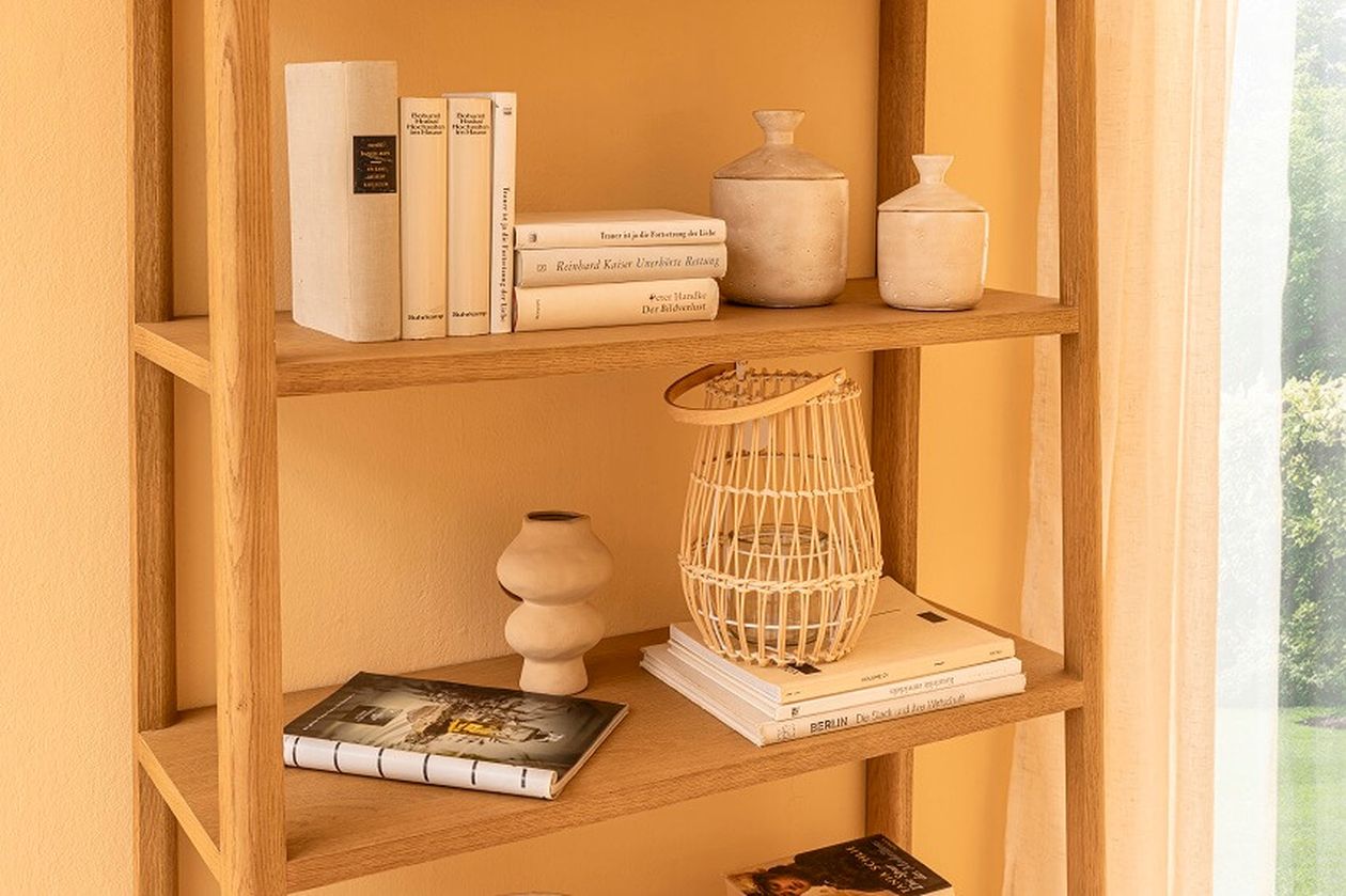 Ein Holzregal mit Büchern, Keramik, einer Weidenlaterne und einer dekorativen Vase in einem sanft beleuchteten Raum.