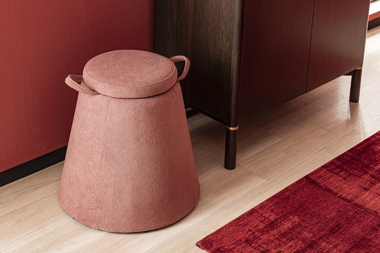 Pinkfarbener, kegelförmiger Hocker mit Griffen neben einem dunklen Holzschrank auf hellem Holzboden, roter Teppich im Vordergrund.