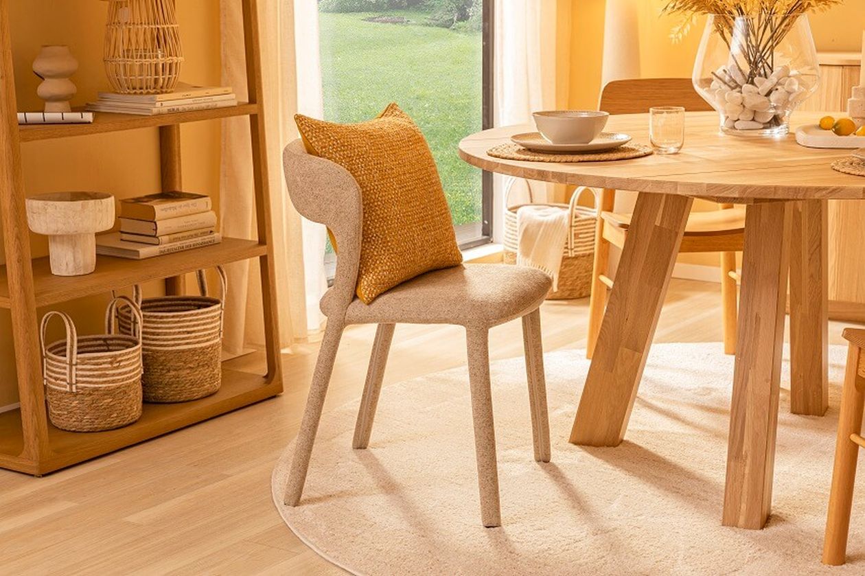 Gemütlicher Essbereich mit rundem Holztisch, Stuhl mit gelbem Kissen, Regal mit Büchern und geflochtenen Körben, in warmen Beige- und Holztönen gehalten.