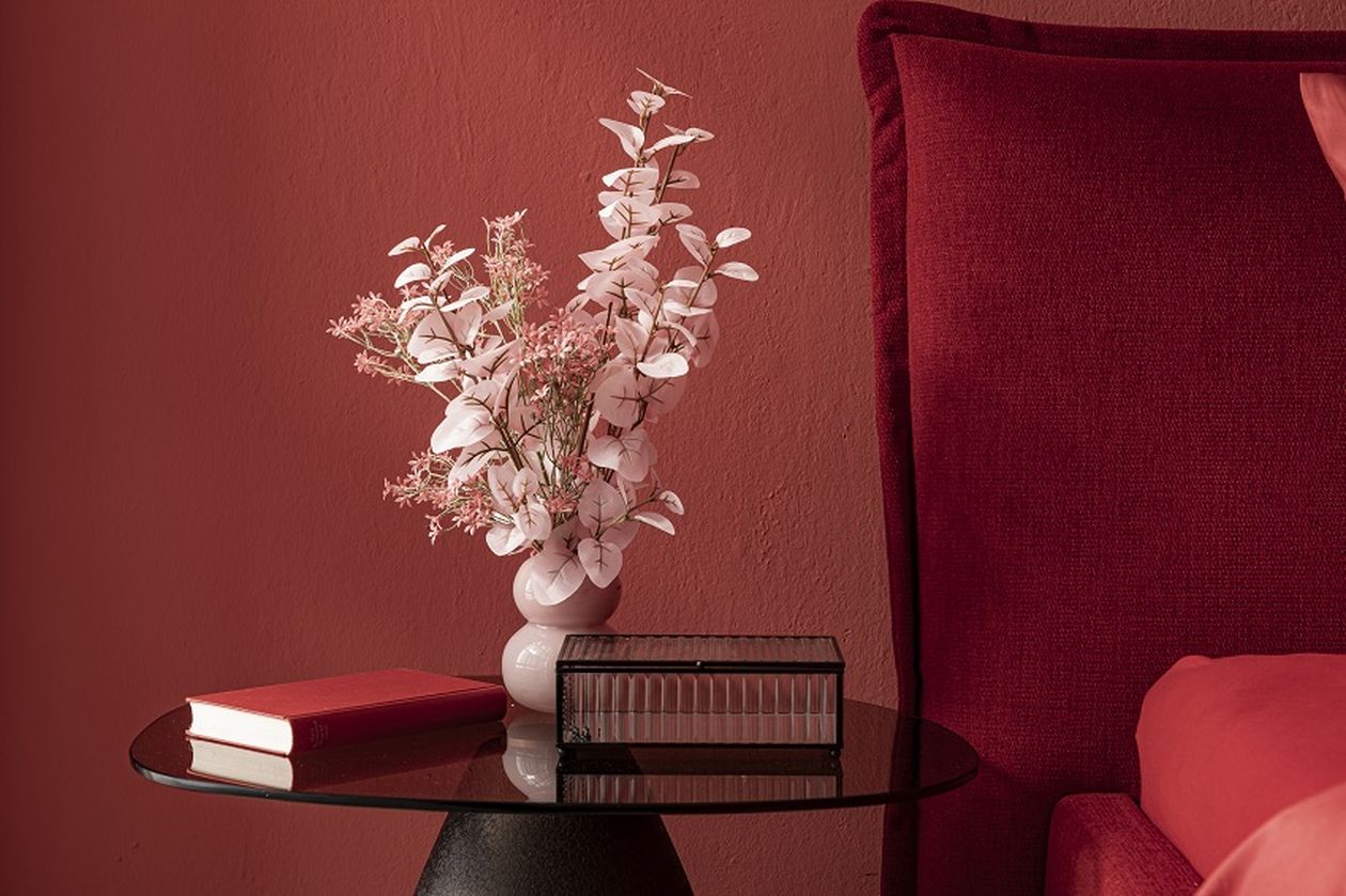 Ein schwarzer Tisch mit einer rosa Vase voller Blumen, einem roten Buch und einer Glasbox, neben einem roten Sofa in einem roten Raum.