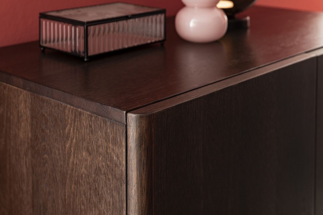Eckbereich eines dunklen Holzschranks, darauf eine geriffelte Glasbox und eine rosa Keramikdekoration, vor einer roten Wand.