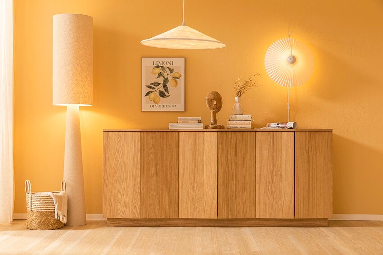Wohnzimmer mit gelber Wand, Stehlampe, Hängelampe, Holzanrichte, Zierobjekten, und einem Zitronenbild. Korb und Bücher dekorieren den Raum.