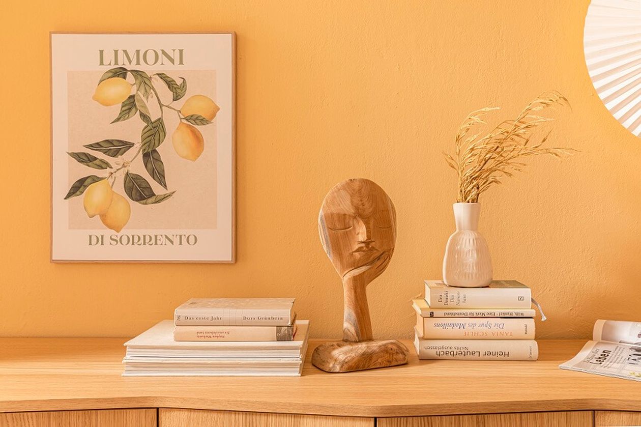 Holzskulptur, Bücher, Vase mit getrocknetem Gras auf Holzkommode vor gelber Wand mit Zitronenbild. Harmonisch und stilvoll arrangiert.