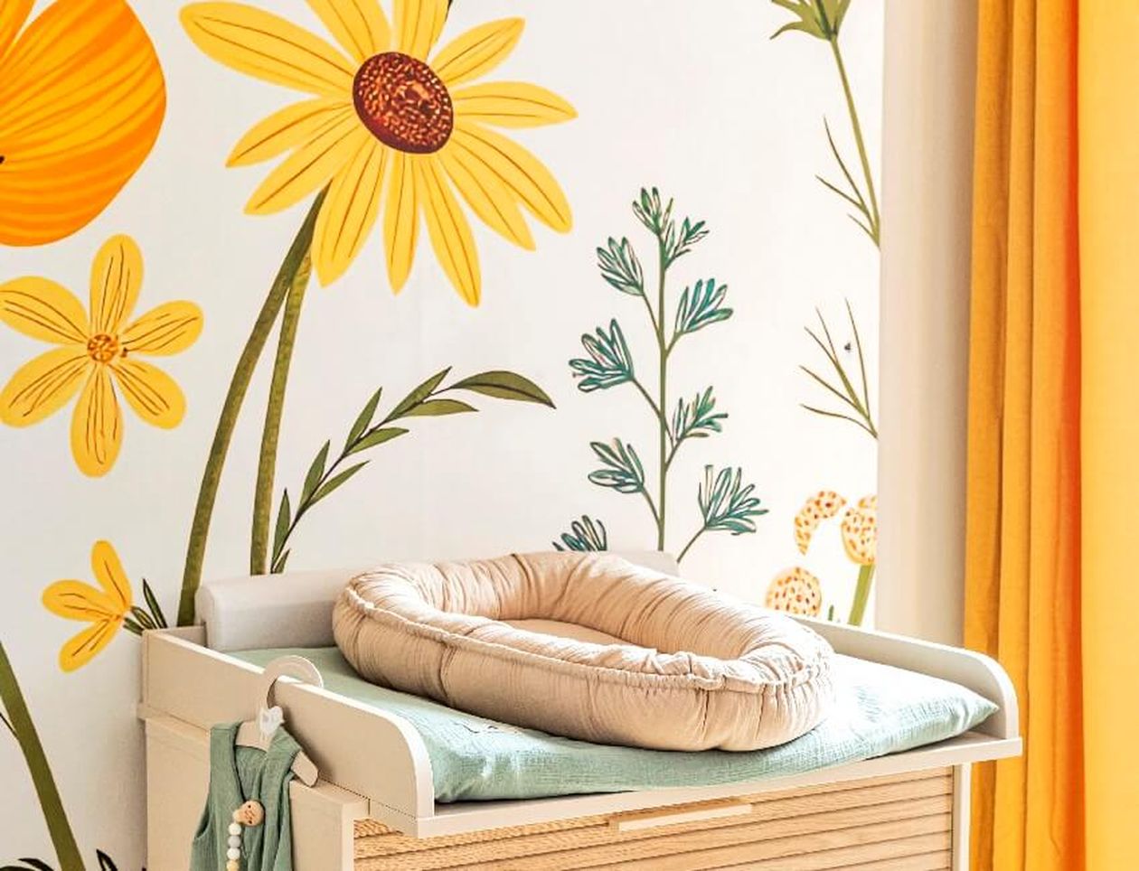 Wickelkommode mit kuscheligen Unterlagen in Beige und Türkis mit Blumentapete an der Wand.