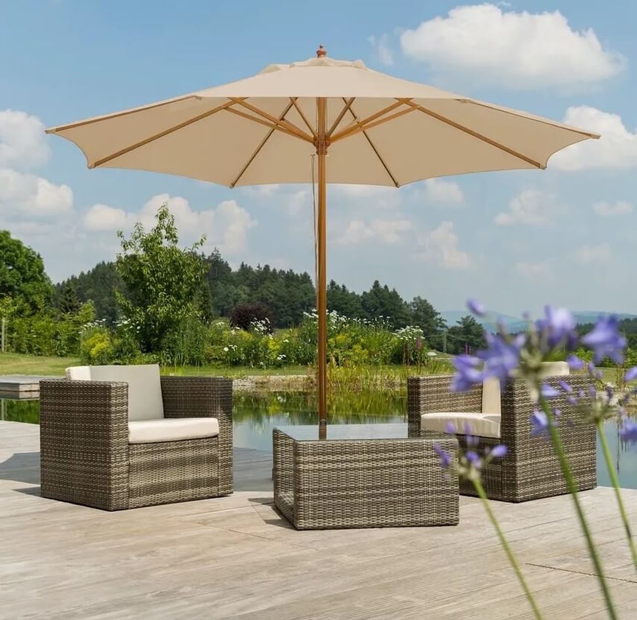 Außenbereich mit Rattanstühlen und einem großen weißen Sonnenschirm auf einer Holzterrasse am Teich, unter einem blauen Himmel mit verstreuten Wolken.