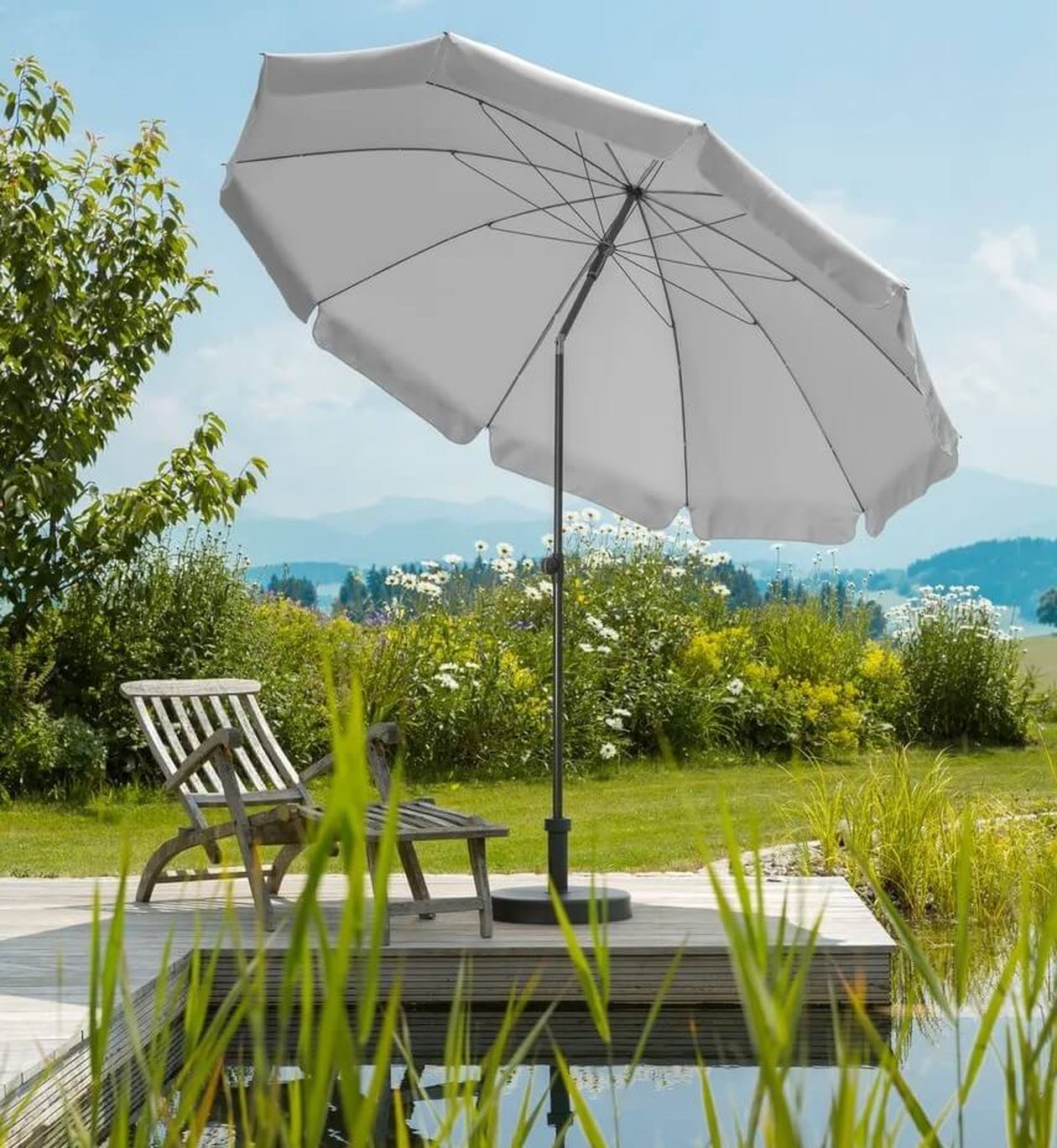 Ein weißer Sonnenschirm spendet Schatten für einen Holzliegestuhl am Teich, umgeben von üppigem Grün und Wildblumen unter einem strahlend blauen Himmel.