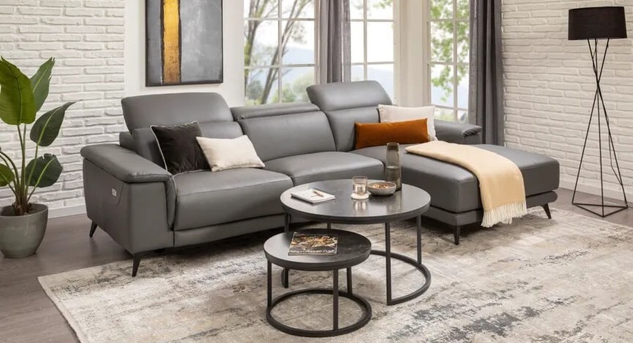 Modernes Wohnzimmer mit einem grauen Sofa, runden Couchtischen, einer Stehlampe und einem großen Fenster. Zur Einrichtung gehören Pflanzen und abstrakte Kunst.