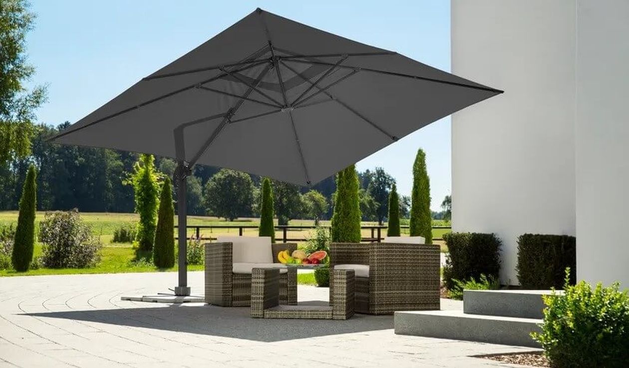 Terrasse mit Rattanmöbeln unter einem großen grauen Sonnenschirm, umgeben von Grün und einem weißen Gebäude, an einem sonnigen Tag.