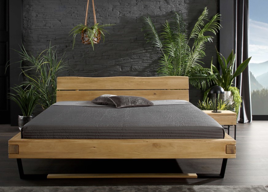 Modernes Schlafzimmer mit einem hölzernen Bettrahmen, grauer Bettwäsche, Topfpflanzen und einer Hängepflanze an einer dunklen Backsteinwand.