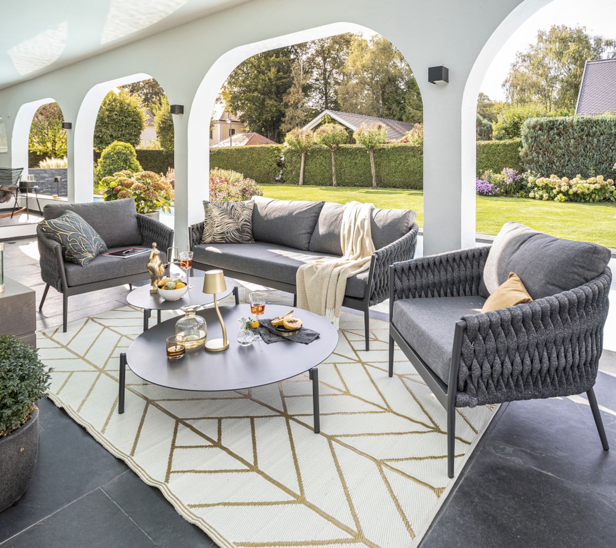 Gemütliche überdachte Terrasse mit grauen Sitzmöbeln, rundem Couchtisch, dekorativen Kissen und Blick auf einen gepflegten Garten.
