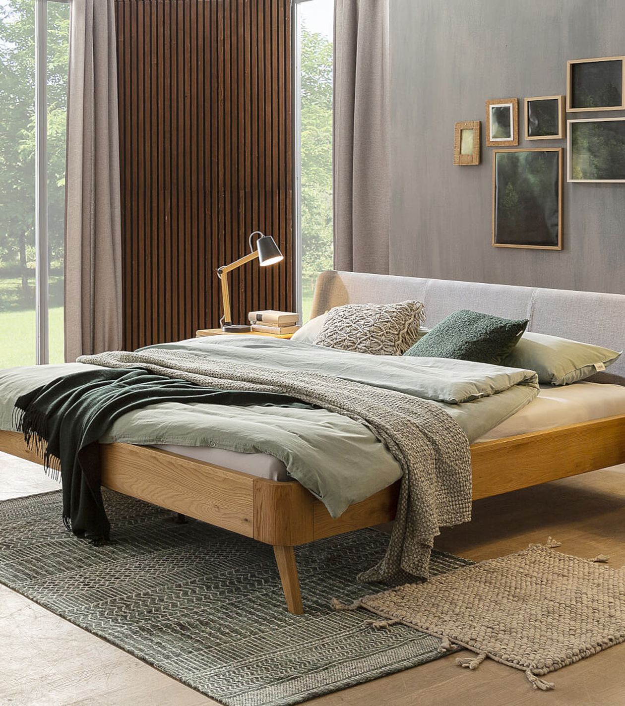 Modernes Schlafzimmer mit einem Holzbett im Mid-Century-Stil, grüner Bettwäsche, strukturierten Überwürfen und einer Lampe auf einem Nachttisch aus Holz. Großes Fenster mit Blick auf den Garten.