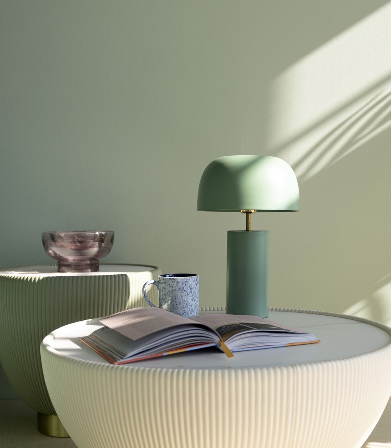 Minimalistischer Tisch mit offener Buchseite, grüner Tischlampe, blauer Kaffeetasse und rosafarbenem Deko-Becken auf gerippten, weißen Beistelltischen.