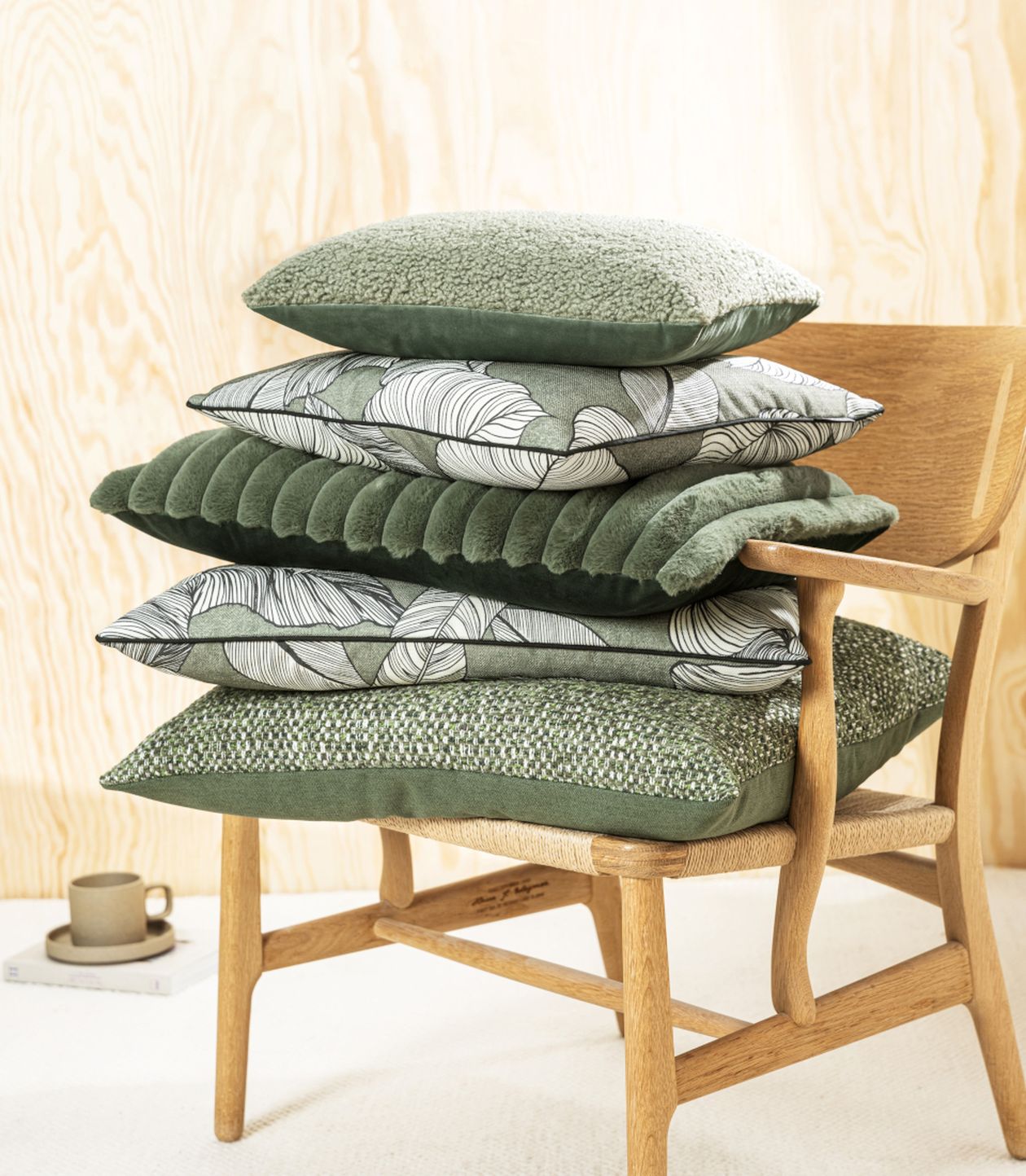 Stapel aus fünf Kissen in verschiedenen Grün- und Beigetönen mit unterschiedlichen Mustern wie Blättern, Samt und Strickoptik. Die Kissen liegen auf einem hellen Holzstuhl mit geflochtenem Sitz.