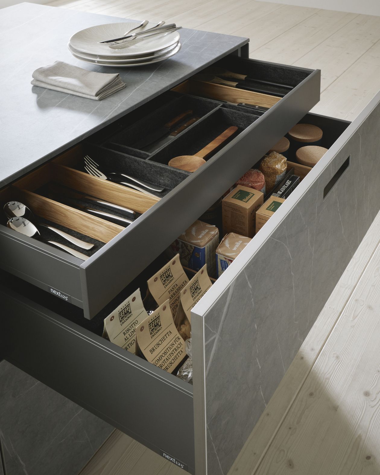 Offene, moderne Küchenschubladen mit ordentlich sortiertem Besteck, Holzlöffeln und verschiedenen verpackten Vorratsartikeln unter einer Steinarbeitsplatte.
