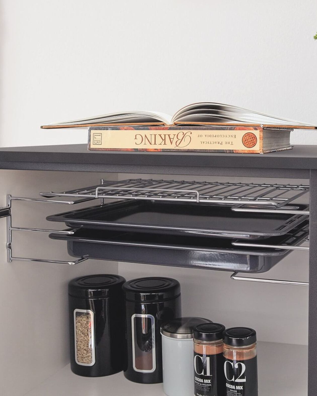 Schrank mit ausziehbaren Ablagen zur Aufbewahrung von Backblechen und einem Kühlgitter, darunter schwarze Vorratsdosen und Gewürzgläser, obenauf ein offenes Backbuch.