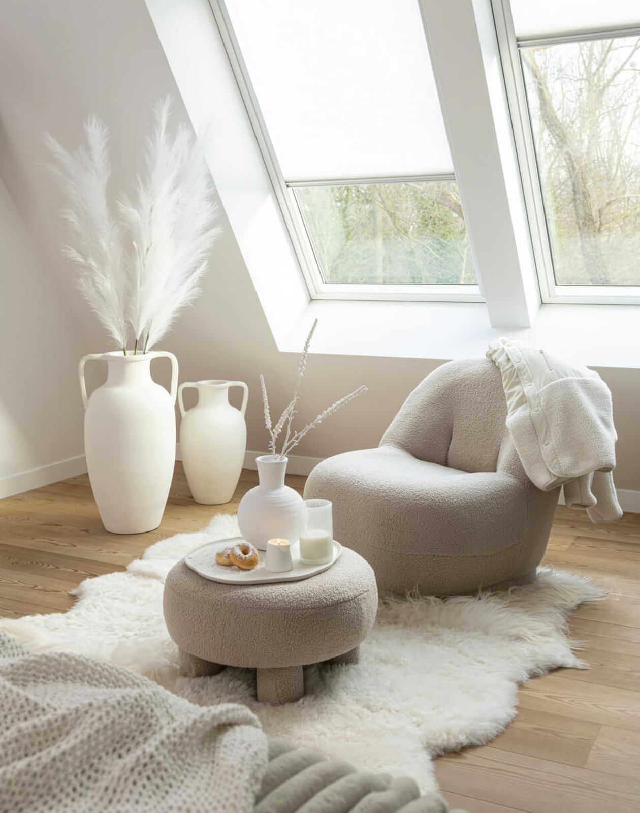 Gemütliches Zimmer mit einem Plüschsessel, einem runden Hocker, weißer Einrichtung und hohen Vasen an großen, schrägen Fenstern. Neutrale Töne und weiche Stoffe.