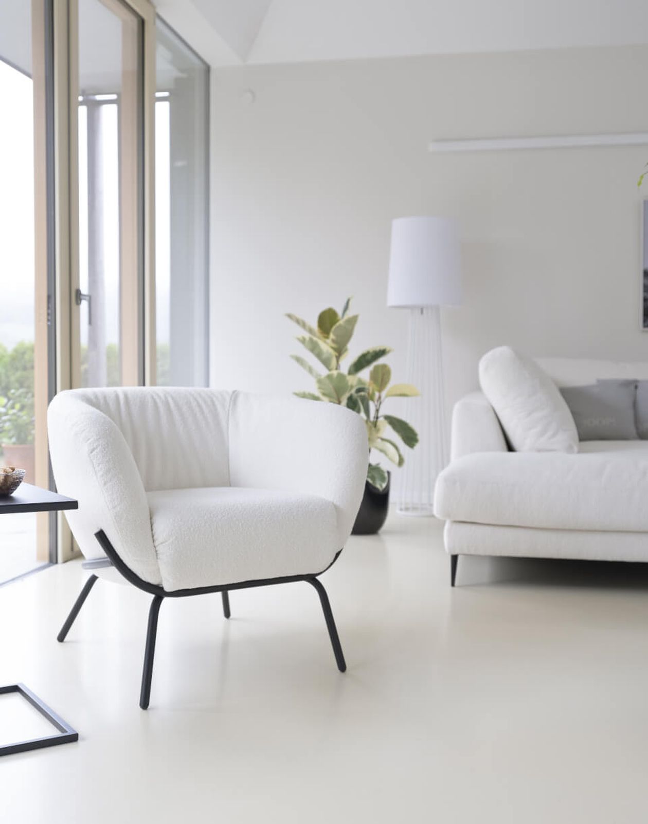Minimalistisches Wohnzimmer mit weißem Sessel, Sofa und Stehlampe. Große Fenster und eine Topfpflanze verstärken den hellen, luftigen Eindruck des Raumes.