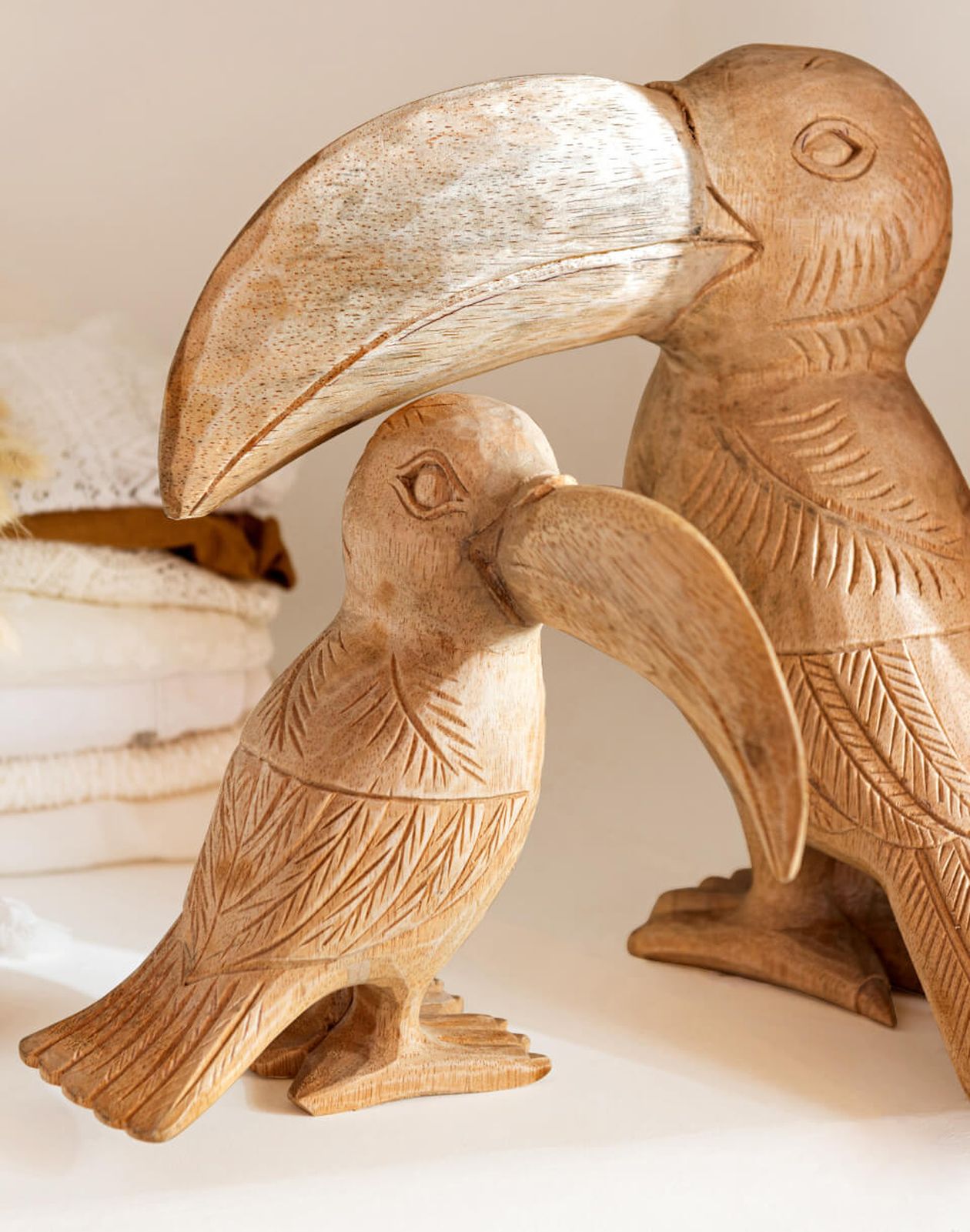 Zwei hölzerne Tukan-Skulpturen mit detaillierten Schnitzereien, eine größer als die andere, stehen auf einer Fläche neben gefaltetem Stoff.