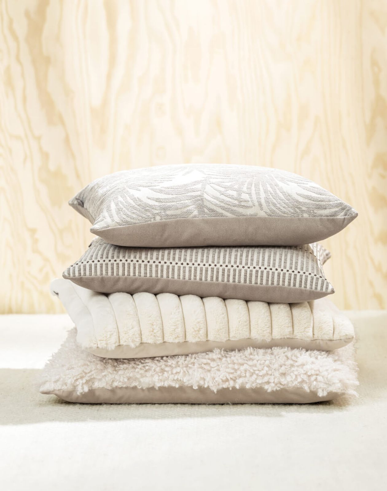 Ein Stapel neutralfarbener Kissen und Decken auf einem weichen, beigen Untergrund vor einem hellen Holzgrund.