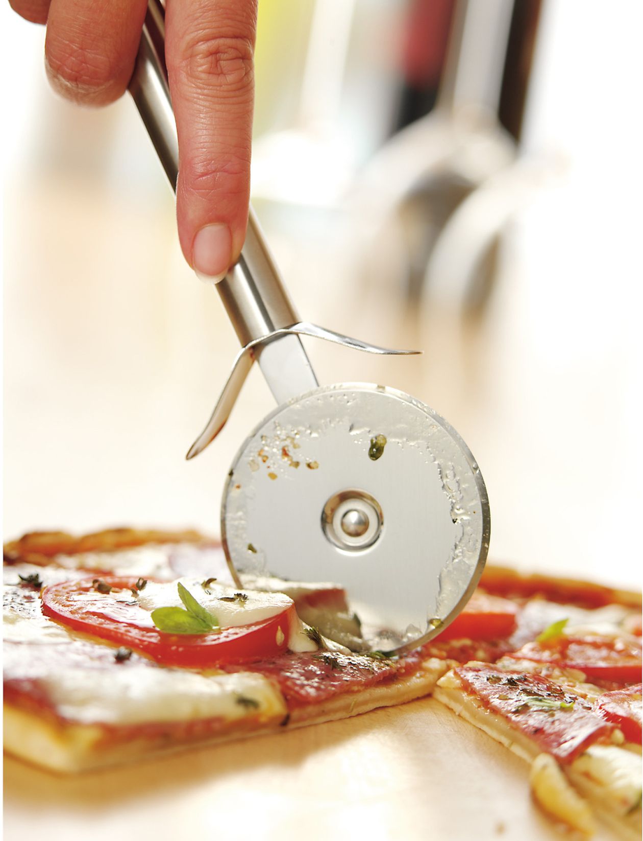 Eine Hand hält einen Pizzaschneider aus Edelstahl und schneidet eine frische Tomaten-Basilikum-Pizza mit geschmolzenem Käse auf einem Holzbrett.