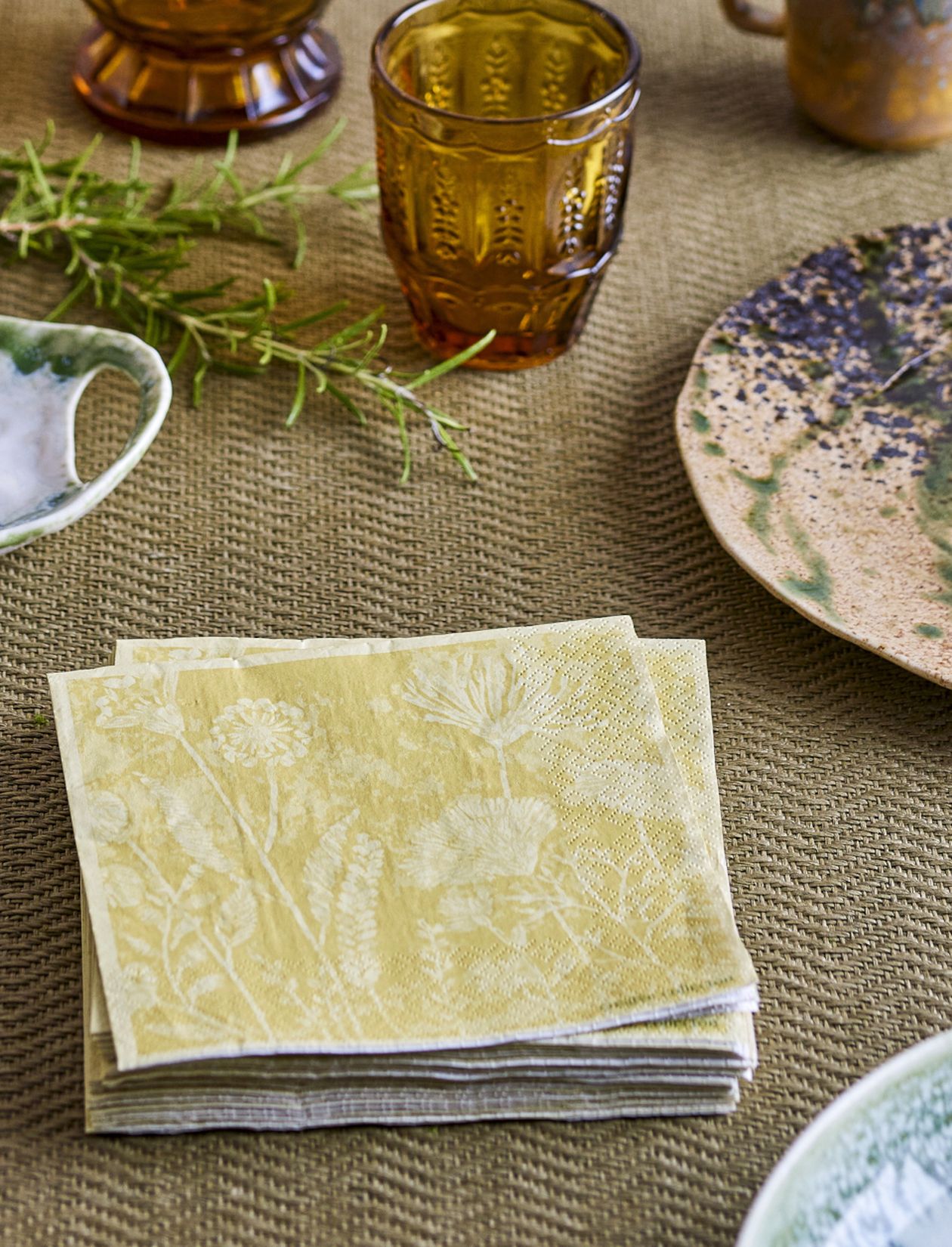 Ein Stapel gelber, blumengemusterter Papierservietten auf einer strukturierten braunen Tischdecke, daneben ein bernsteinfarbenes Glas, ein Keramikteller und ein Rosmarinzweig.