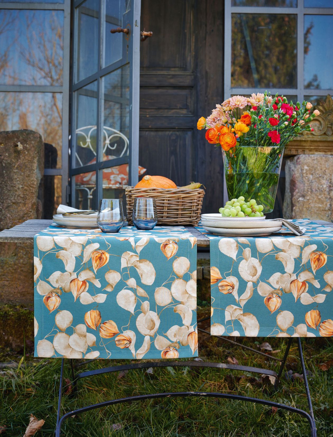 Rustikaler Gartentisch mit blauen Blumenläufern, Vase mit orangefarbenen und rosafarbenen Blumen, Korb mit Brot, Tellern, Gläsern und Weintrauben neben einer Holztür.
