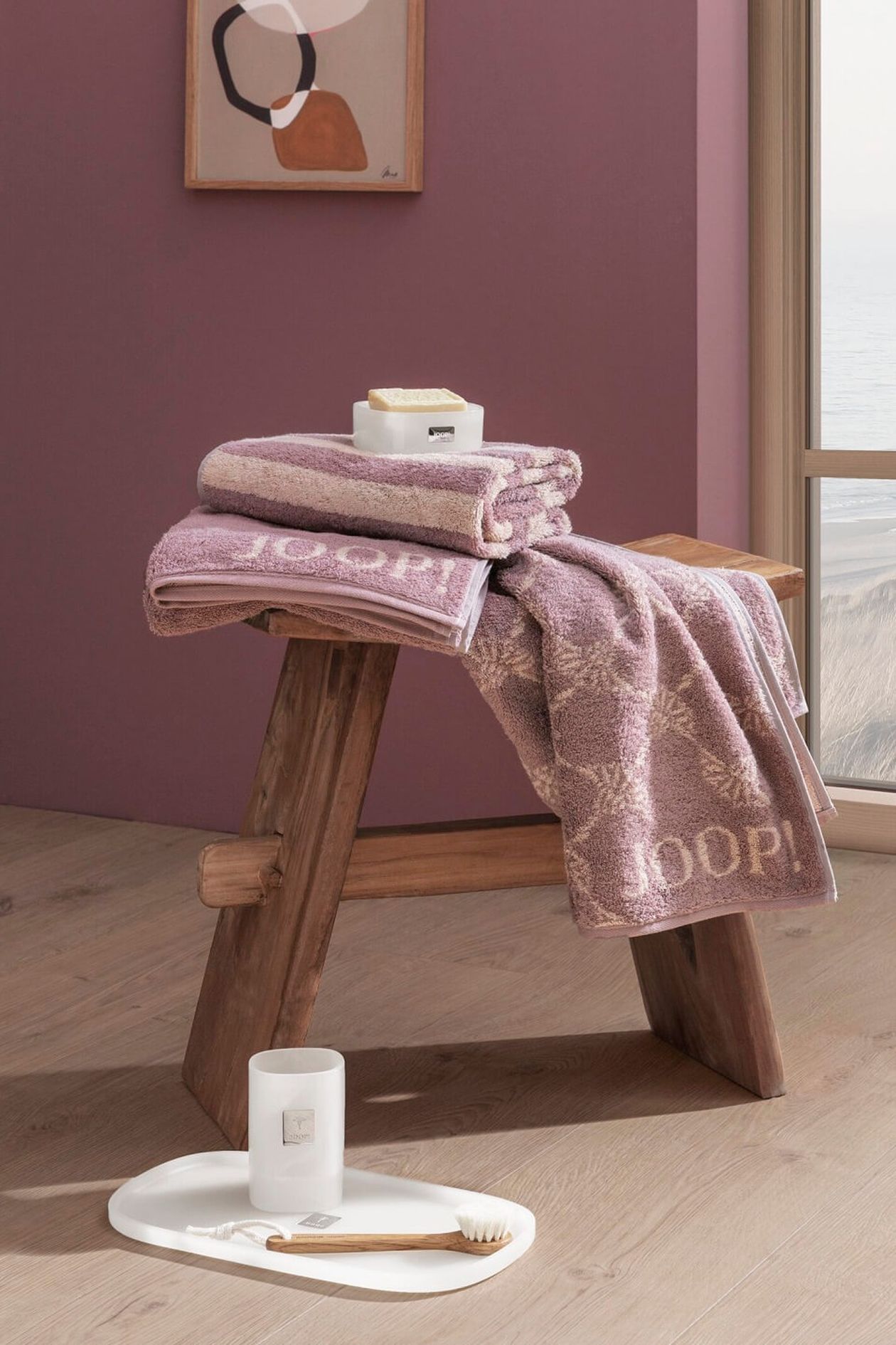 In einem gemütlichen Raum mit rosa Wänden liegt eine rustikale Holzbank mit gefalteten lila Handtüchern. Daneben steht ein Tablett mit einer Kerze und einer Bürste auf dem Boden.