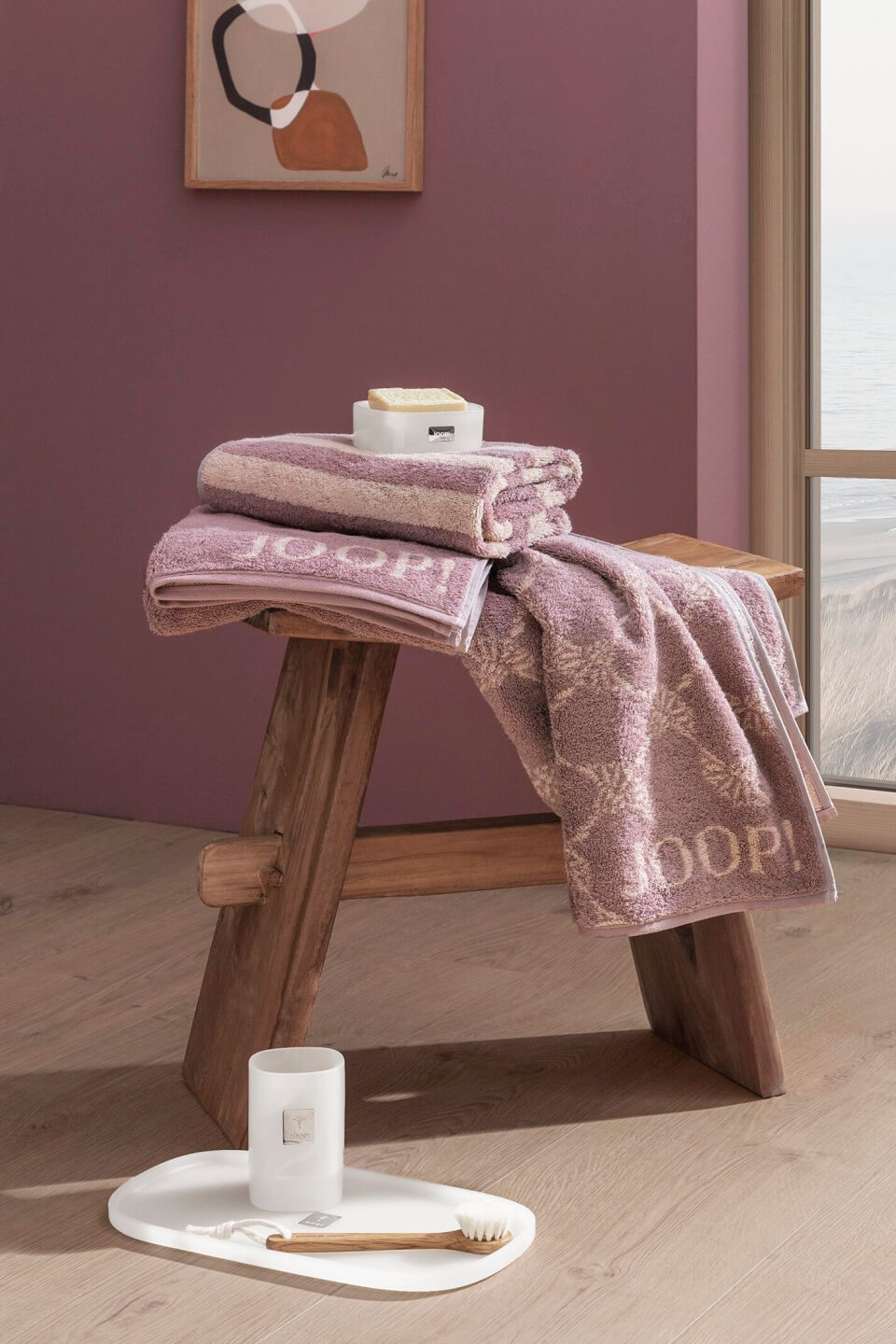 In einem gemütlichen Raum mit rosa Wänden liegt eine rustikale Holzbank mit gefalteten lila Handtüchern. Daneben steht ein Tablett mit einer Kerze und einer Bürste auf dem Boden.