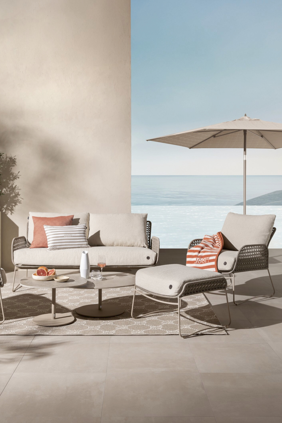 Auf einer hellen, mediterranen Terrasse steht eine Gruppe beiger Loungemöbel mit Apricot-Weiß gestreiften Outdoordecken und -kissen der Serie Essence von JOOP!.
