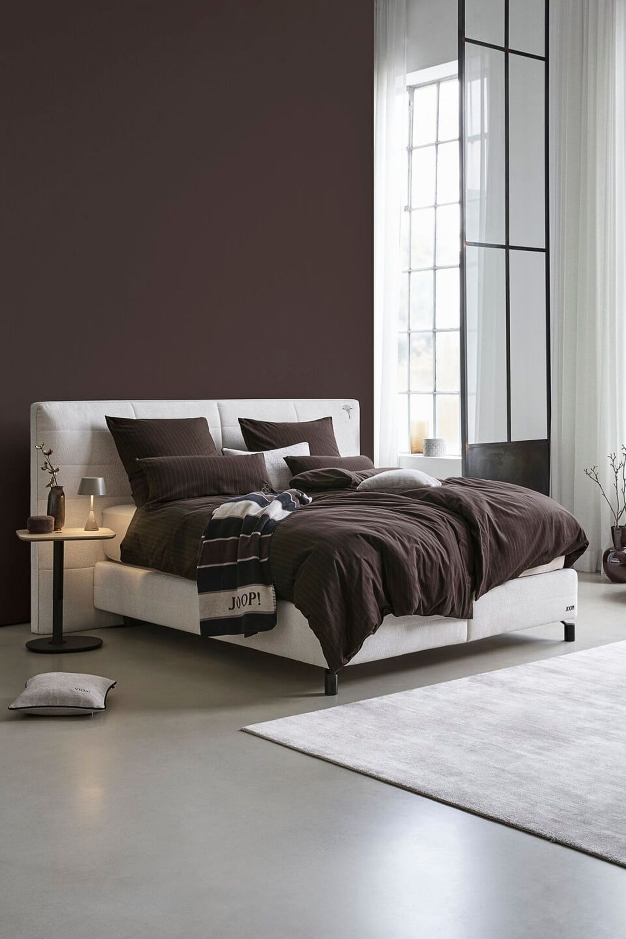 Modernes Schlafzimmer mit weißem Bett, brauner Bettwäsche und Beistelltisch. Großes Fenster und dunkle Akzentwand schaffen eine gemütliche Atmosphäre.